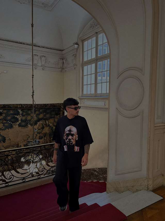 Remera negra Thairos bajando una escalera, junto a un tapiz antiguo.